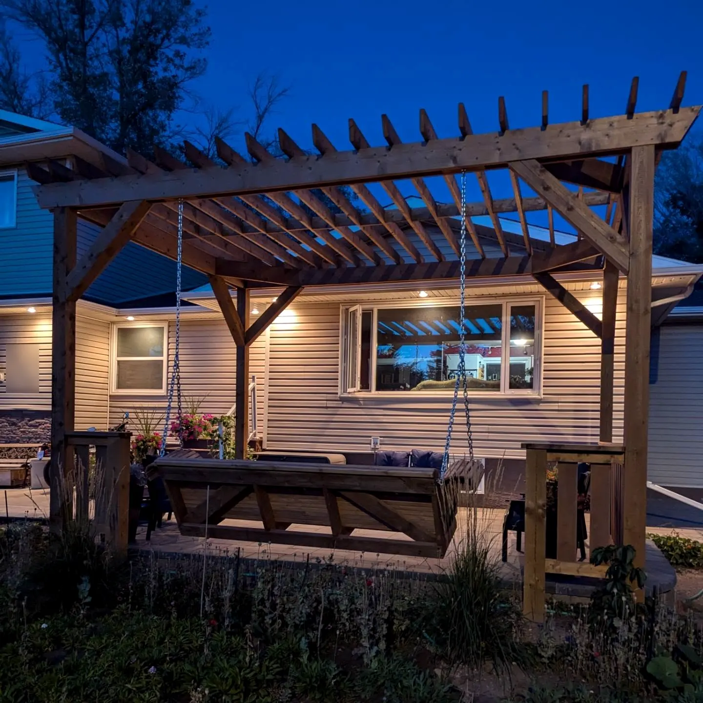 Timber pergola with hanging bench swing at dusk, lit house behind
